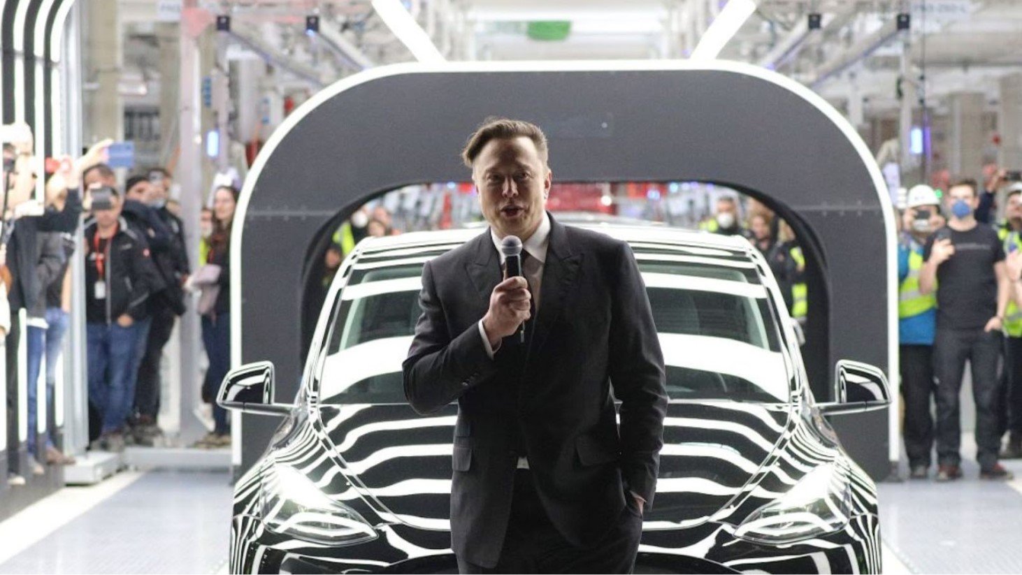 Elon Musk in a suit holding a microphone speaking in a factory setting, with a car and audience members in the background