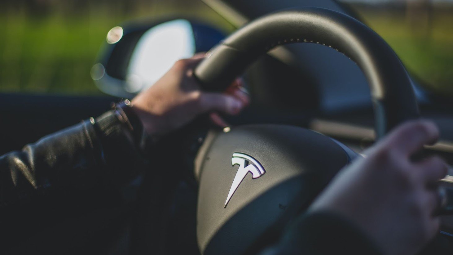 Close-up of a hand on a Tesla steering wheel, focusing on the Tesla logo, with a blurred background of the car's side mirror
